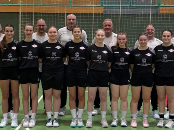 Eine Mädchen-Sportmannschaft in schwarzen Uniformen steht in einer Turnhalle mit ihren Trainern, die beigefarbene Hemden tragen. Die Gruppe posiert gemeinsam auf einem Hallenplatz, mit einem Netz und gelben Sitzen im Hintergrund.