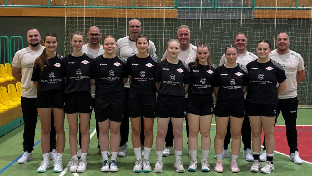 Eine Mädchen-Sportmannschaft in schwarzen Uniformen steht in einer Turnhalle mit ihren Trainern, die beigefarbene Hemden tragen. Die Gruppe posiert gemeinsam auf einem Hallenplatz, mit einem Netz und gelben Sitzen im Hintergrund.