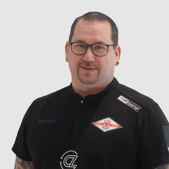 Ein Mann mit kurzen dunklen Haaren, Brille und Spitzbart trägt ein schwarzes Handball-Brandenburg-Shirt mit Vereinslogos und Sponsorenaufnähern. Er steht vor einem schlichten hellgrauen Hintergrund, schaut in die Kamera und lächelt leicht.