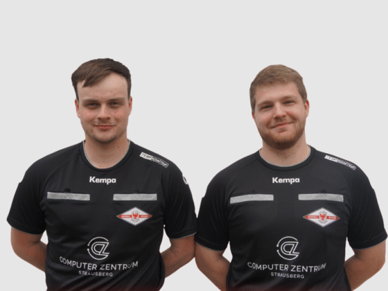 Zwei Männer in schwarzen Sporttrikots mit Sponsorenlogos und einem rot-weißen Emblem stehen nebeneinander und lächeln leicht in die Kamera. Ihre Kleidung lässt vermuten, dass es sich um Schiedsrichter handelt, die vor einem schlichten weißen Hintergrund posieren.