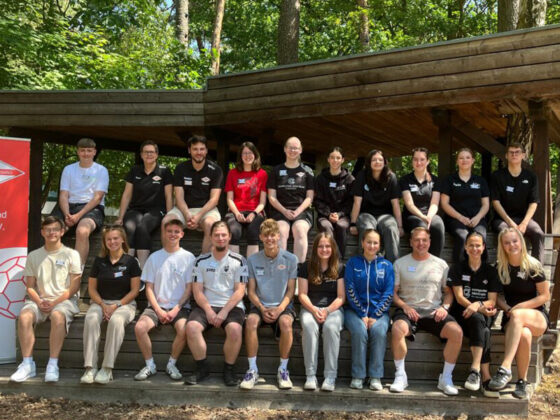 Eine Gruppe von 20 jungen Erwachsenen sitzt und steht draußen auf Holzstufen zusammen und posiert für ein Gruppenfoto. Sie tragen Freizeit- oder Sportkleidung und unterstreichen damit ihren Jugendarbeit-Handball-Geist. Links ist eine rot-weiße Fahne des Handball-Verbandes Brandenburg zu sehen.
