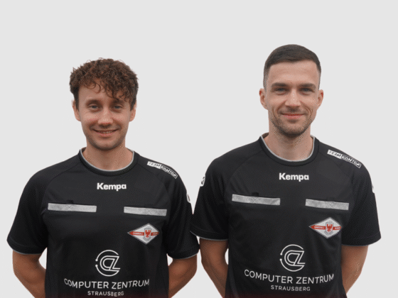 Zwei junge Männer in passenden schwarzen Sporttrikots mit Sponsorenlogos, Kempa-Branding und Schiedsrichter-Handball-Abzeichen stehen nebeneinander vor einem schlichten hellen Hintergrund und lächeln in die Kamera.