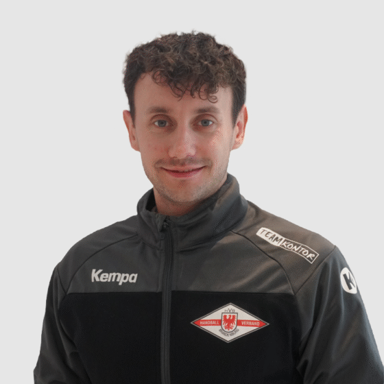 Ein Mann mit kurzen lockigen braunen Haaren, der eine schwarz-graue Handball Brandenburg Sportjacke mit Logos trägt, steht vor einem schlichten hellen Hintergrund und lächelt in die Kamera.