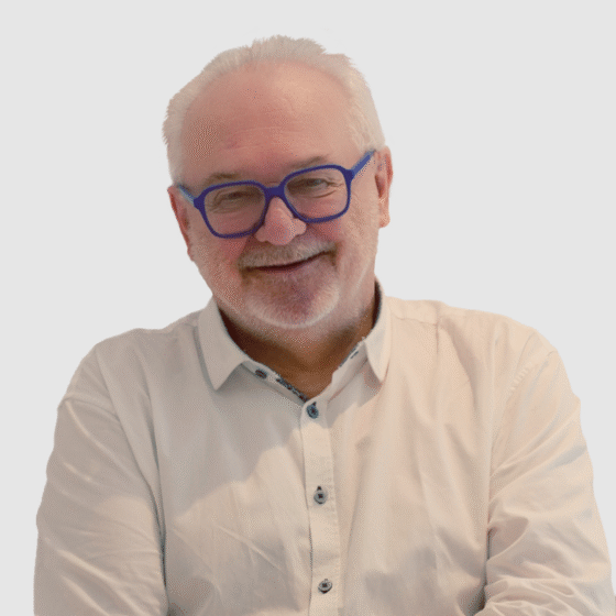 Ein älterer Mann mit weißen Haaren, Bart und blauer Brille lächelt freundlich. Er trägt ein helles Button-up-Hemd und steht vor einem schlichten Hintergrund - vielleicht schwelgt er in Erinnerungen an seine Zeit bei Handball Brandenburg.