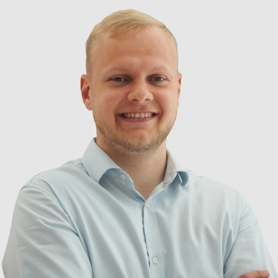 Ein Mann mit kurzen blonden Haaren und einem Bart lächelt, trägt ein hellblaues Hemd mit Kragen und steht mit verschränkten Armen vor einem schlichten weißen Hintergrund.