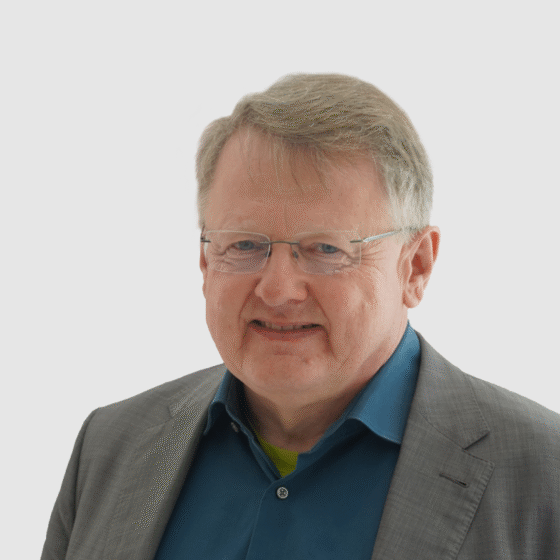 Ein Mann mit hellem Haar und Brille, der ein blaues Hemd und einen grauen Blazer trägt und vor einem schlichten weißen Hintergrund lächelt, verkörpert den Geist von Handball Brandenburg.