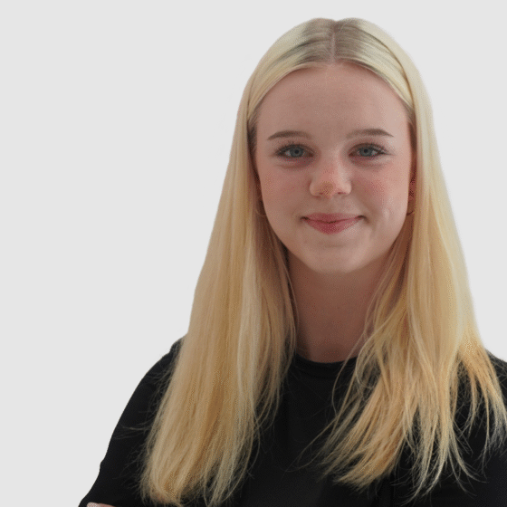 Eine junge Frau mit langen blonden Haaren und einem schwarzen Hemd lächelt sanft vor einem schlichten hellgrauen Hintergrund.