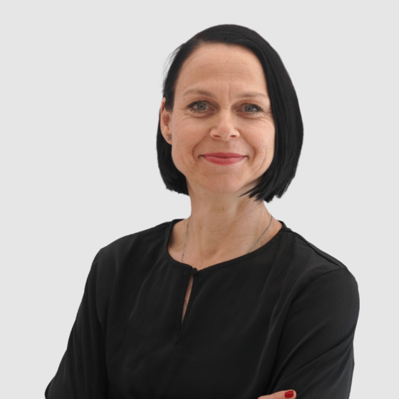 Eine Frau mit glattem schwarzem Haar, die ein schwarzes langärmeliges Oberteil trägt, steht mit verschränkten Armen und lächelt selbstbewusst vor einem schlichten hellgrauen Hintergrund.