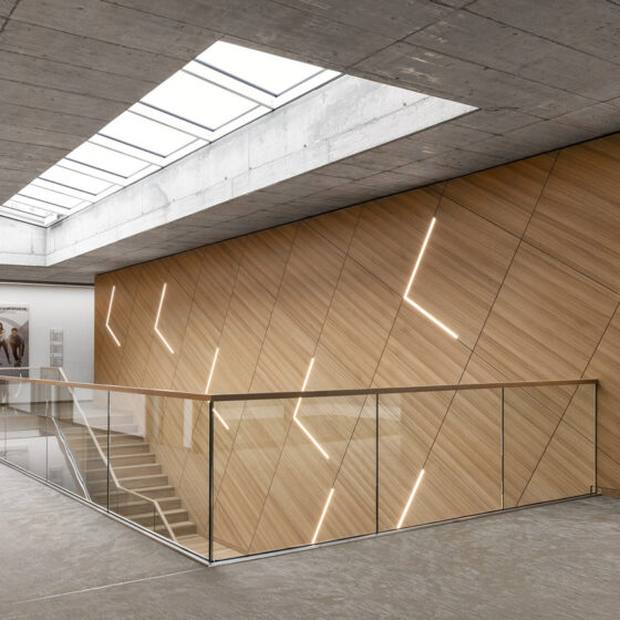 Ein moderner Flur mit einem Oberlicht, einer Betondecke und holzgetäfelten Wänden mit diagonalen LED-Lichtleisten. Ein Glasgeländer umgibt eine Treppe, und an der weißen Wand am Ende des Flurs hängt gerahmte Kunst.