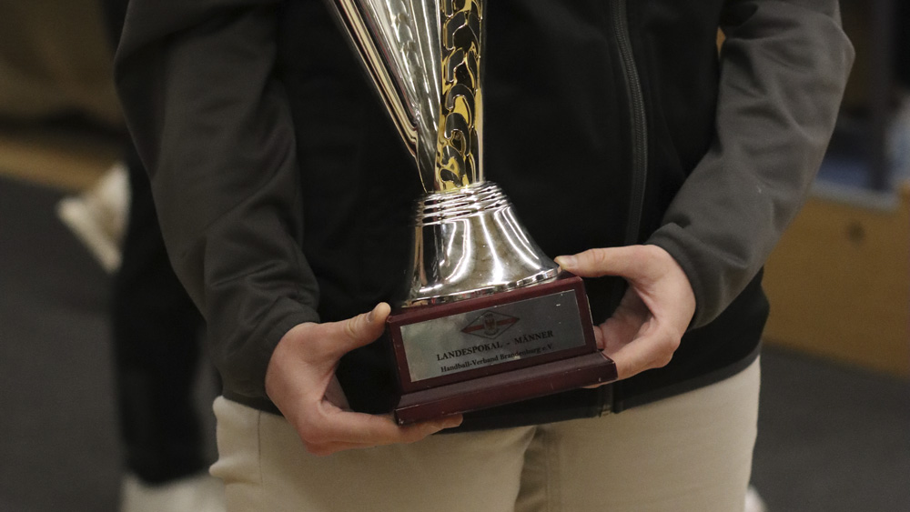 Eine Person in schwarzer Jacke und beiger Hose hält eine große silberne Trophäe mit einem Holzsockel und einer eingravierten Plakette. Der Text auf der Plakette ist teilweise sichtbar, aber nicht ganz klar.