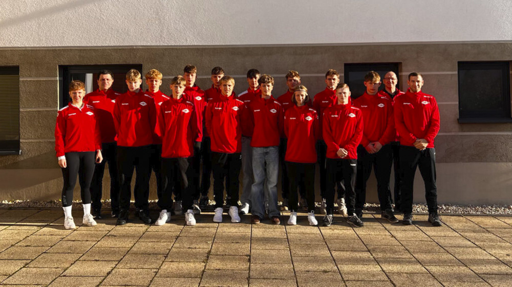 Eine Gruppe von fünfzehn Personen steht draußen auf einer gepflasterten Fläche und posiert für ein Foto. Sie alle tragen passende leuchtend rote Jacken und schwarze Hosen und scheinen Teil eines Teams oder einer Organisation zu sein.