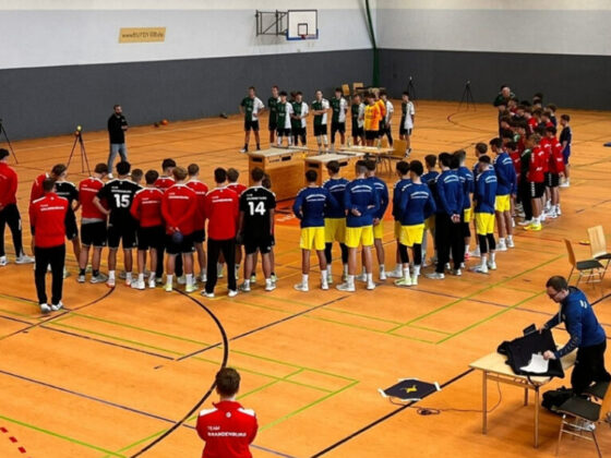 Eine Gruppe von Sportteams in farbenfrohen Uniformen steht in einer Sporthalle und bildet einen großen Kreis um einen Tisch, anscheinend während einer Preisverleihung oder Zeremonie. Einige Offizielle und Trainer sind in der Nähe, mit Stühlen und Ausrüstung, die um den Raum herum aufgebaut sind.
