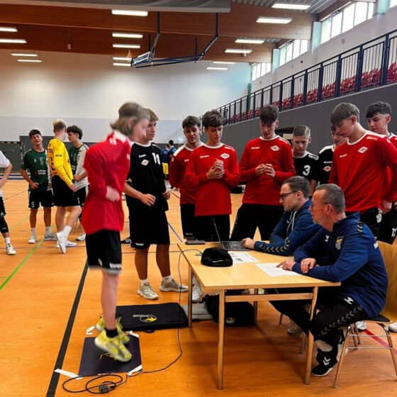 Eine Gruppe Jugendlicher in Sportuniformen sieht zu, wie ein Junge in einem roten Hemd während eines Fitnesstests auf einer Matte springt, während zwei Erwachsene an einem Tisch in einer Sporthalle die Ergebnisse aufzeichnen.