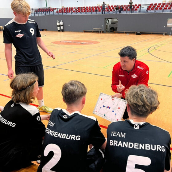 Ein Trainer in einem roten Trainingsanzug kniet auf dem Boden einer Sporthalle und erklärt vier jungen Sportlern in schwarzen "Team Brandenburg"-Uniformen während eines Trainings oder Spiels in der Halle Strategien auf einer Tafel.