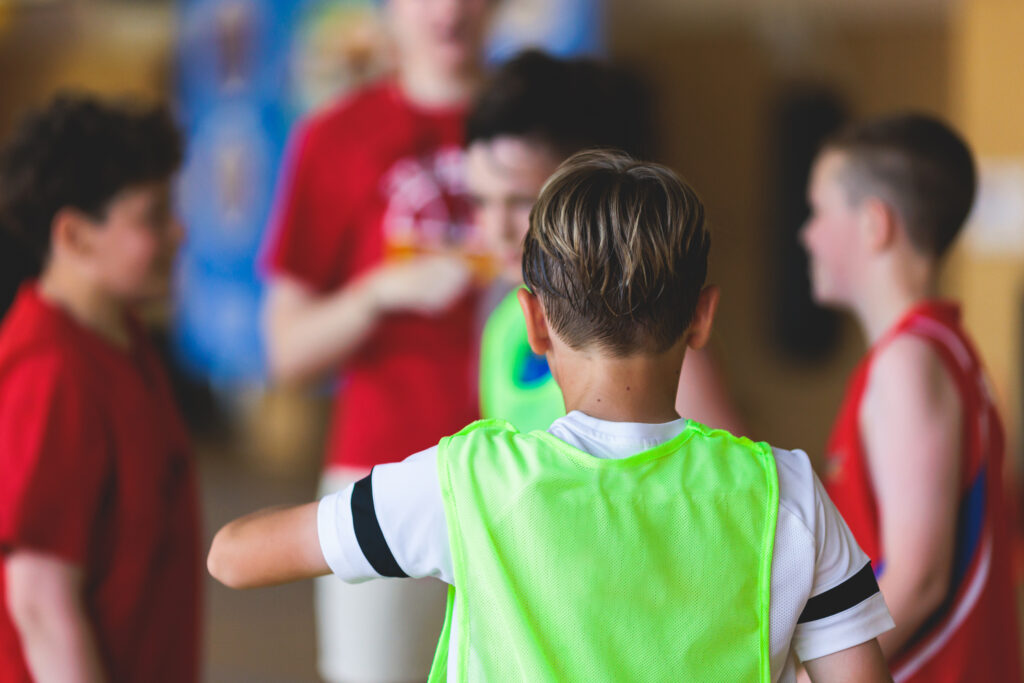 Eine Gruppe von Kindern in Sportkleidung versammelt sich drinnen, wobei ein Kind in einer grünen Weste den Blick abwendet und in Richtung anderer Kinder in roten Hemden gestikuliert, was auf eine Teambesprechung oder eine Spielaktivität hindeutet.