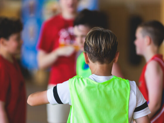 Eine Gruppe von Kindern in Sportkleidung versammelt sich drinnen, wobei ein Kind in einer grünen Weste den Blick abwendet und in Richtung anderer Kinder in roten Hemden gestikuliert, was auf eine Teambesprechung oder eine Spielaktivität hindeutet.