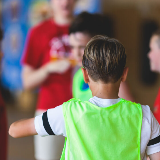 Eine Gruppe von Kindern in Sportkleidung versammelt sich drinnen, wobei ein Kind in einer grünen Weste den Blick abwendet und in Richtung anderer Kinder in roten Hemden gestikuliert, was auf eine Teambesprechung oder eine Spielaktivität hindeutet.