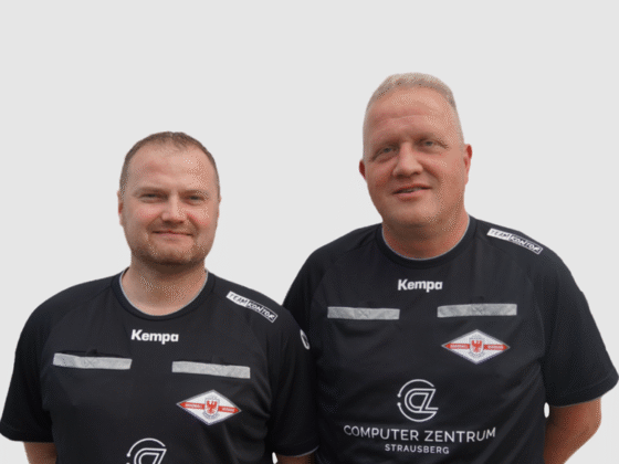 Zwei Männer in passenden schwarzen Schiedsrichter-Handball-Trikots mit Kempa-Branding und rot-weißem Logo stehen nebeneinander und lächeln vor einem schlichten hellgrauen Hintergrund.