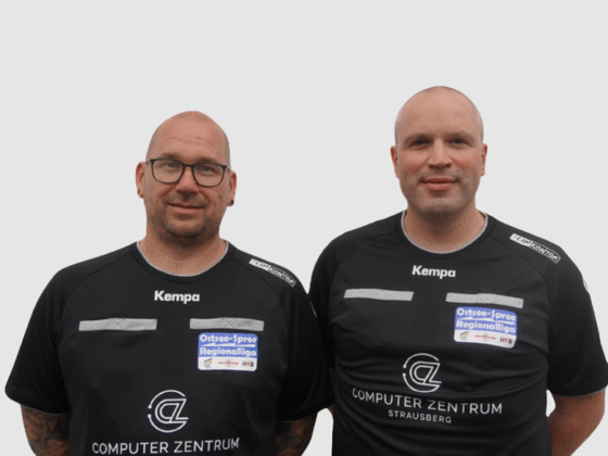 Zwei Männer in schwarzen Sporttrikots mit Logos und Text stehen nebeneinander vor einem schlichten, hellen Hintergrund. Beide haben rasierte Köpfe und Brillen, was darauf hindeutet, dass es sich bei ihnen um Schiedsrichter des Handballvereins oder um Mitglieder einer verwandten Organisation handelt.