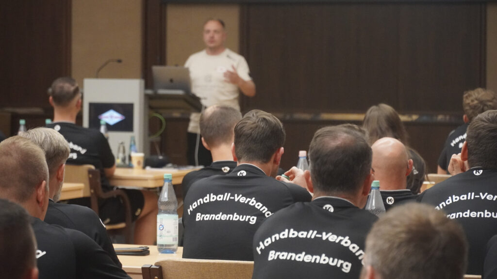 Eine Gruppe von Personen in Shirts des Handball-Verbandes Brandenburg" sitzt in einem Klassenzimmer und hört aufmerksam einem Referenten zu, der vorne an einem Podium und einem Laptop sitzt.