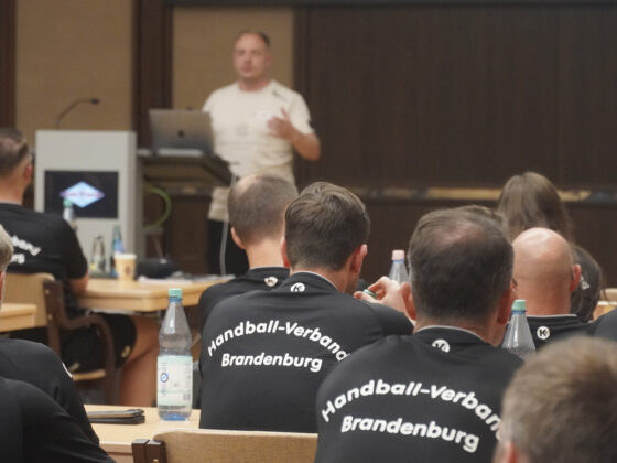 Eine Gruppe von Personen in Shirts des Handball-Verbandes Brandenburg" sitzt in einem Klassenzimmer und hört aufmerksam einem Referenten zu, der vorne an einem Podium und einem Laptop sitzt.