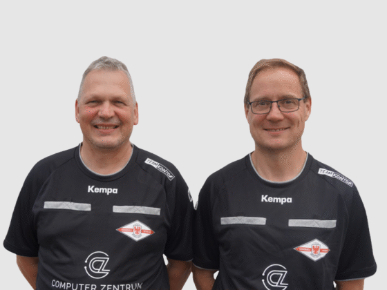Zwei Männer in passenden schwarzen Trikots mit Logos und dem Schriftzug Kempa stehen nebeneinander und lächeln als Schiedsrichter Handball vor einem schlichten weißen Hintergrund in die Kamera.