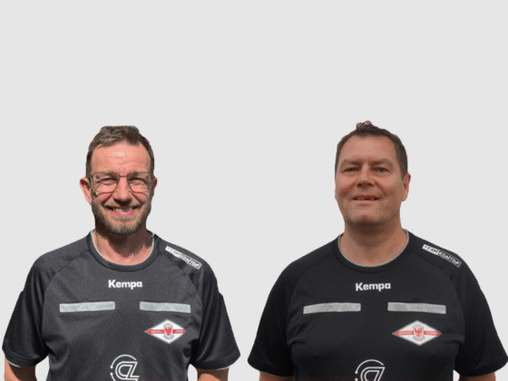 Zwei Männer in schwarzen Sporthemden mit weißem und rotem Logo stehen nebeneinander vor einem schlichten hellgrauen Hintergrund. Beide blicken nach vorne und lächeln, um das professionelle Bild der Handball-Schiedsrichter zu vermitteln.
