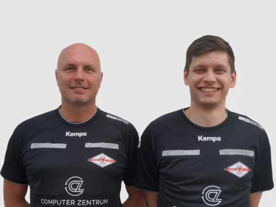 Zwei Handball-Schiedsrichter in passenden schwarzen Kempa-Sportuniformen mit weiß-rotem Wappen stehen nebeneinander und lächeln vor einem schlichten hellen Hintergrund.