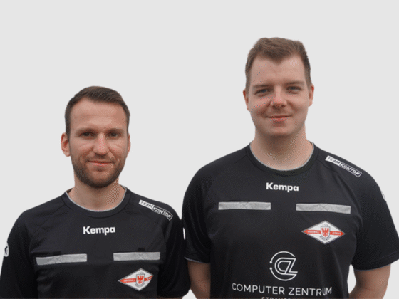 Zwei Männer in schwarzen Schiedsrichter-Handball-Trikots mit weißem und rotem Logo stehen nebeneinander und lächeln in die Kamera vor einem schlichten hellgrauen Hintergrund.