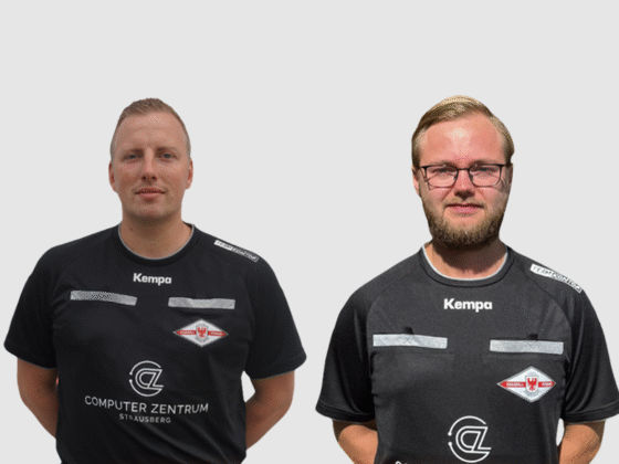 Zwei nebeneinander stehende Männer, beide in schwarzen Sporttrikots mit Kempa-Branding und Vereinslogo auf der Brust, repräsentieren Schiedsrichter Handball vor einem schlichten hellen Hintergrund.