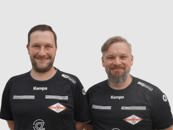 Zwei Männer in schwarzen Kempa-Sportshirts mit Schiedsrichter-Handball-Logos stehen nebeneinander und lächeln in die Kamera vor einem schlichten weißen Hintergrund.
