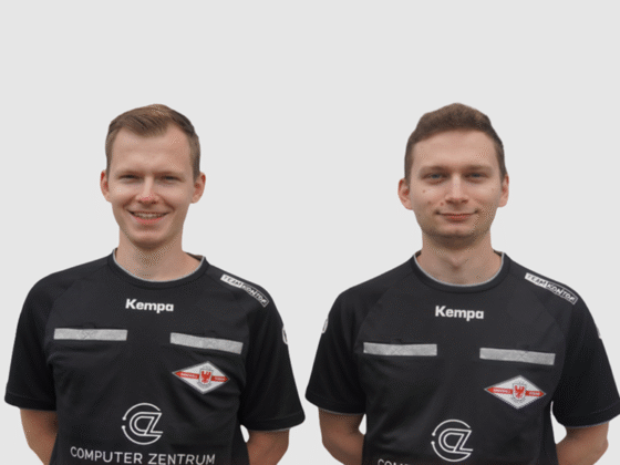 Zwei Männer in schwarzen Sporttrikots mit weißem und rotem Logo, möglicherweise Schiedsrichter im Handball, stehen nebeneinander, schauen nach vorne und lächeln. Der Hintergrund ist schlicht und hellgrau.