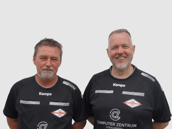 Zwei Männer, Schiedsrichter Handball, stehen Seite an Seite in schwarzen Kempa-Sportshirts mit Logo. Sie lächeln und posieren vor einem schlichten, hellen Hintergrund.