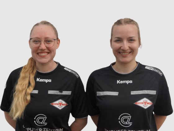 Zwei Frauen mit langen blonden Haaren tragen schwarze Sporttrikots mit Logos, Kempa-Schriftzug und Schiedsrichter-Handball-Abzeichen. Sie stehen nebeneinander und lächeln vor einem schlichten weißen Hintergrund.