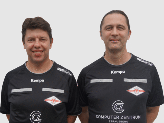 Zwei Männer stehen nebeneinander, beide tragen schwarze Sporthemden mit Kempa und verschiedenen Logos wie Computer Zentrum Strausberg. Die beiden Schiedsrichter Handball lächeln vor einem schlichten hellen Hintergrund.