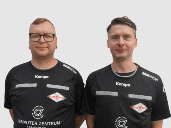 Zwei Männer in schwarzen Sporttrikots mit dem Logo von Schiedsrichter Handball stehen nebeneinander vor einem schlichten, hellen Hintergrund und schauen in die Kamera. Einer trägt eine Brille und hat kurze Haare, der andere hat einen ähnlichen Haarschnitt und keine Brille.
