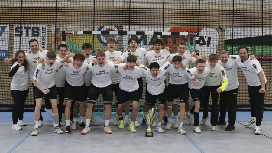 Eine Gruppe junger Männer und ihre Trainer in passenden weißen "Super Sächsencup"-Shirts feiern drinnen hinter einer großen Trophäe, lächelnd und jubelnd, die Arme umeinander gelegt.