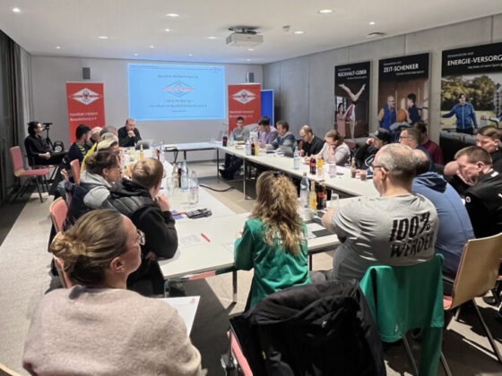 Eine Gruppe von Personen sitzt um einen großen, U-förmigen Konferenztisch in einem modernen Besprechungsraum und hört einem Redner an der Stirnseite zu. Eine Präsentation wird an die Wand projiziert, und im Raum sind verschiedene Banner angebracht.