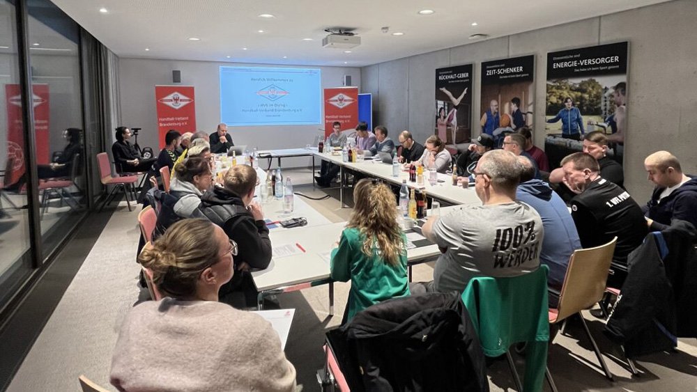 Eine Gruppe von Personen sitzt um einen großen, U-förmigen Konferenztisch in einem modernen Besprechungsraum und hört einem Redner an der Stirnseite zu. Eine Präsentation wird an die Wand projiziert, und im Raum sind verschiedene Banner angebracht.