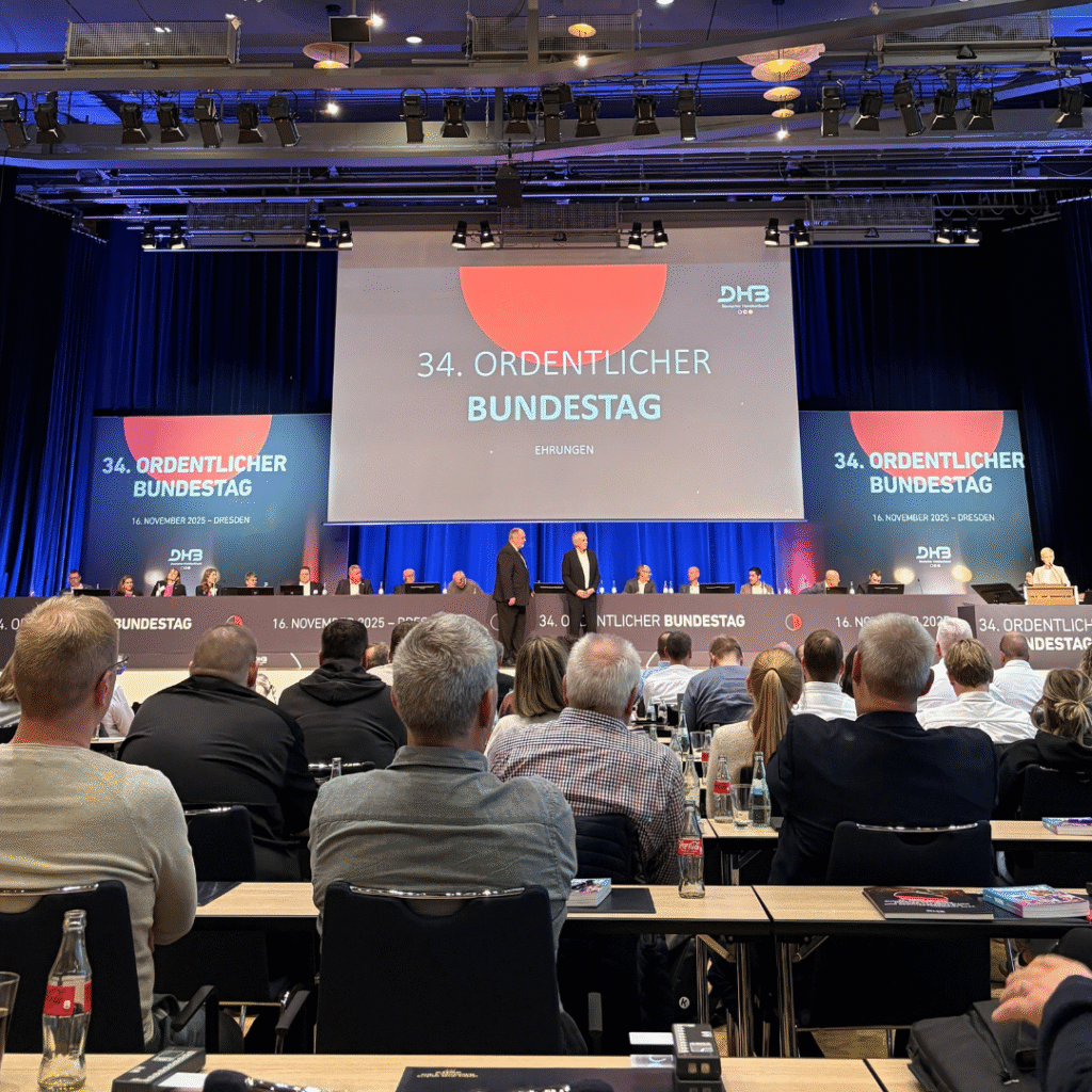 Ein Konferenzsaal mit einem Publikum, das auf eine Bühne blickt, auf der zwei Personen vor einem großen Bildschirm stehen, auf dem "34. Ordentlicher Bundestag". Der Rahmen ist formell, mit Tischen, Mikrofonen und blau-roter Beleuchtung.