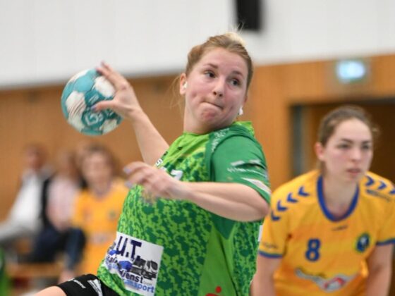 Eine Handballspielerin in einem grünen Trikot bereitet sich darauf vor, den Ball während eines Spiels zu werfen, während eine andere Spielerin in einem gelben Trikot von hinten zusieht. Das Spiel findet in einer Halle statt, mit unscharfen Zuschauern im Hintergrund.