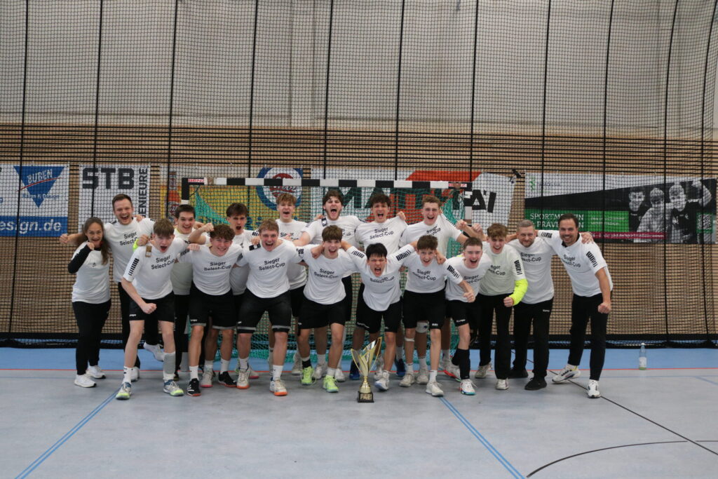 Eine jubelnde Hallenfußballmannschaft in passenden weißen Hemden und schwarzen Shorts posiert mit einer Trophäe und feiert ihren Sieg in einer Sporthalle mit Fahnen im Hintergrund.