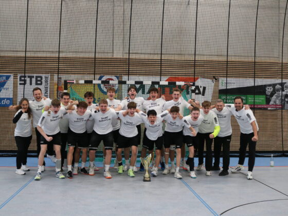 Eine jubelnde Hallenfußballmannschaft in passenden weißen Hemden und schwarzen Shorts posiert mit einer Trophäe und feiert ihren Sieg in einer Sporthalle mit Fahnen im Hintergrund.