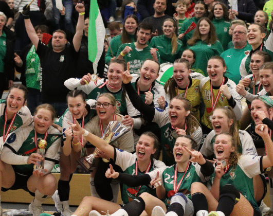 Eine jubelnde Frauenhandballmannschaft und ihre Fans feiern gemeinsam in einer Halle, lächelnd und jubelnd, einige halten einen Pokal und Medaillen. In der begeisterten Menge hinter ihnen werden grüne und weiße Fahnen geschwenkt.