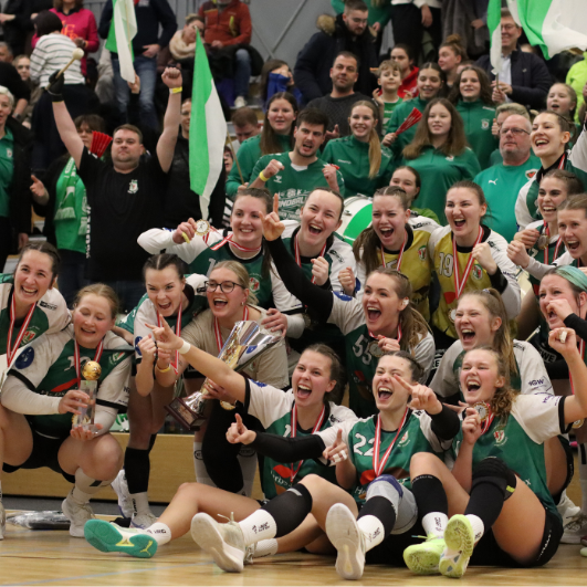 Eine jubelnde Frauenhandballmannschaft und ihre Fans feiern gemeinsam in einer Halle, lächelnd und jubelnd, einige halten einen Pokal und Medaillen. In der begeisterten Menge hinter ihnen werden grüne und weiße Fahnen geschwenkt.