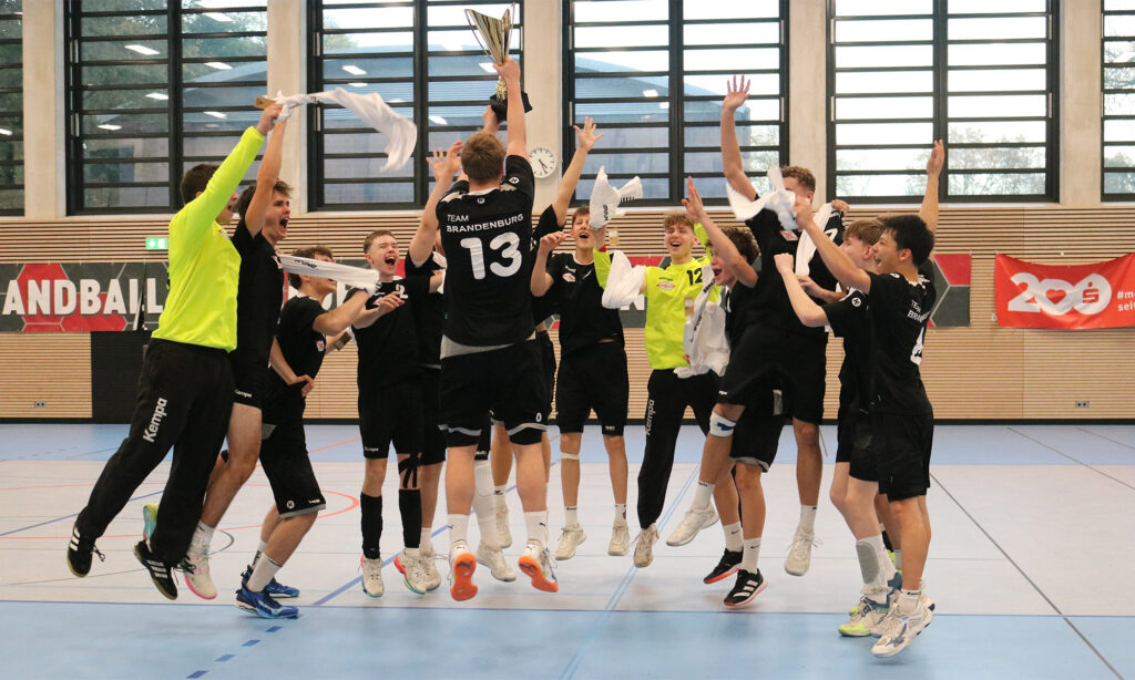 Eine fröhliche Männerhandballmannschaft in schwarzen Trikots springt und feiert gemeinsam in der Halle, während ein Spieler einen Pokal hochhält. Ganz im Sinne des Handball-Leistungssports winken einige Spieler mit Handtüchern und alle lächeln vor Begeisterung.
