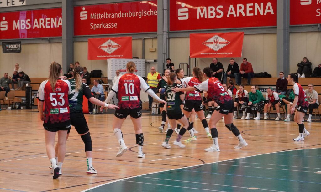 Ein Handballspiel der Frauen in einer Sporthalle ist im Gange. Spielerinnen in roten und schwarzen Trikots verteidigen sich gegen Gegnerinnen in Schwarz und Grün. Zuschauer und Mannschaftsbetreuer sitzen auf Bänken an der Seite. Darüber hängen Banner für die MBS ARENA.