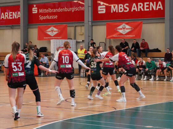 Ein Handballspiel der Frauen in einer Sporthalle ist im Gange. Spielerinnen in roten und schwarzen Trikots verteidigen sich gegen Gegnerinnen in Schwarz und Grün. Zuschauer und Mannschaftsbetreuer sitzen auf Bänken an der Seite. Darüber hängen Banner für die MBS ARENA.