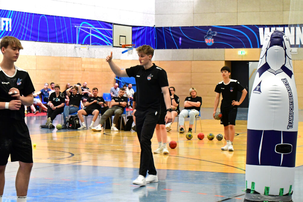 Ein Trainer zeigt die Daumen nach oben, während er junge männliche Sportler in einer Turnhalle während einer Handball-Trainingseinheit anleitet, wobei Bälle und eine Übungspuppe zu sehen sind. Im Hintergrund sind Zuschauer zu sehen.