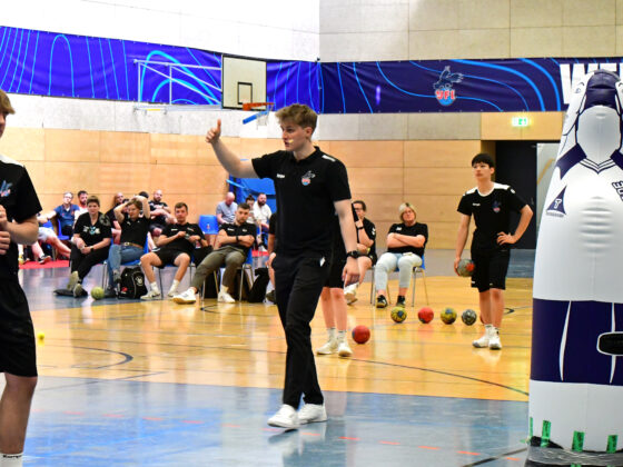 Ein Trainer zeigt die Daumen nach oben, während er junge männliche Sportler in einer Turnhalle während einer Handball-Trainingseinheit anleitet, wobei Bälle und eine Übungspuppe zu sehen sind. Im Hintergrund sind Zuschauer zu sehen.